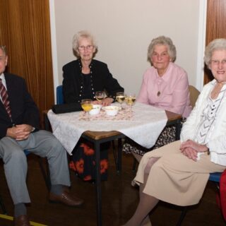 Four older adults seated around a lace-covered table at a social gathering.