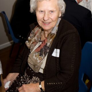 Joan seated on a blue chair at a social event, smiling.