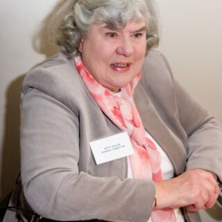 Individual seated with a mobility aid, wearing a light gray blazer and floral pink scarf.