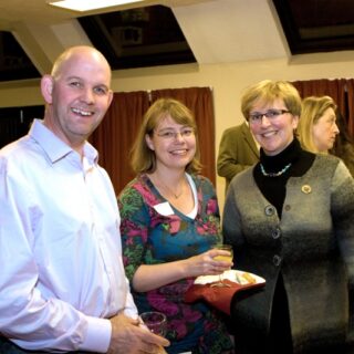 Three attendees in the foreground, smiling and holding drinks and food.
