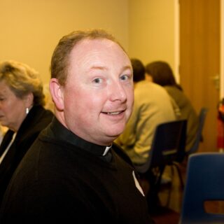 Clergy member seated at a community event, looking toward the camera.