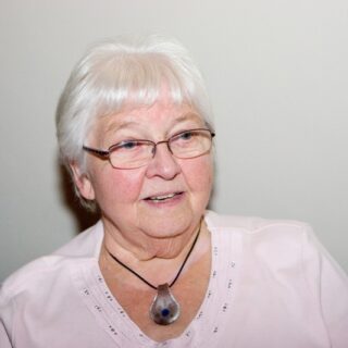Individual with short white hair and glasses, wearing a light pink shirt and pendant necklace.