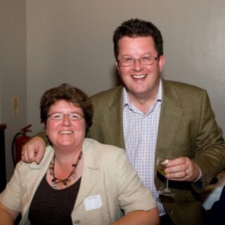 Man and woman with name badges smiling at the anniversary event; the man holds a glass of wine.