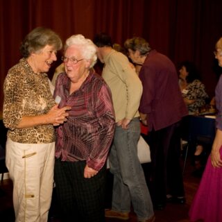 Two women chatting and smiling at a community gathering, with other attendees in the background.