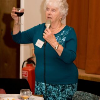 Margaret giving a toast with a microphone and a glass of wine during the Basinga’s 35th anniversary celebration.