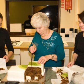 Three women cutting up the celebration cake in the kitchen.