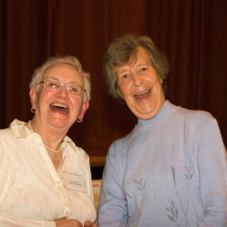 Two women smile at a community event; one wears a U3A committee name badge.